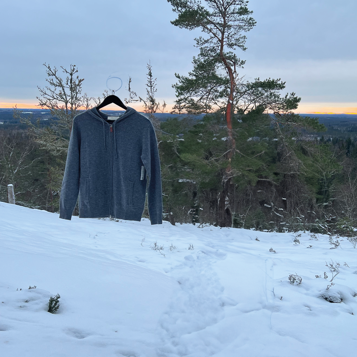 En mörkgrå kashmir zip hoodie med slät textur och en mjuk glans. Tröjan har en fullängds dragkedja framtill, långa ärmar, och en bekväm huva. Den är designad för både stil och värme, perfekt för en kylig dag. Hoodiens kvalitativa kashmirtyg ger en lyxig känsla mot huden.